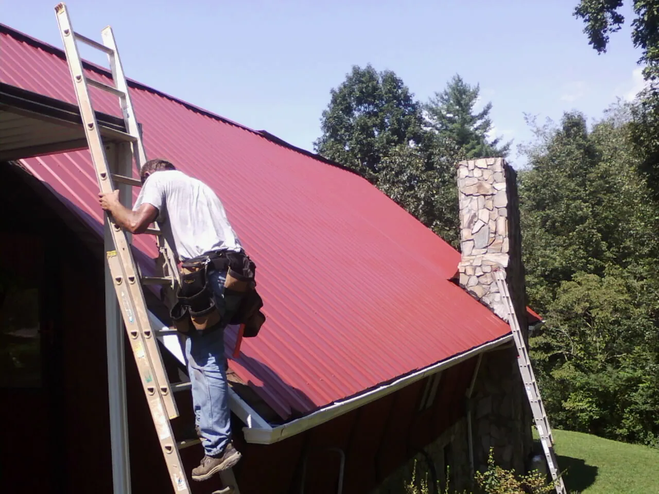 Roofer on a ladder beside a red metal roof and stone chimney — Soffit & Fascia Repair by Roofing Brothers in Oviedo